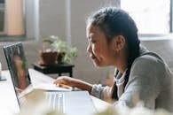 Side view of smart Asian girl sitting at desk and watching laptop in light room